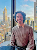 Donny Badamo posing next to a window with the New York City skyline in the background, wearing a striped shirt with curly hair